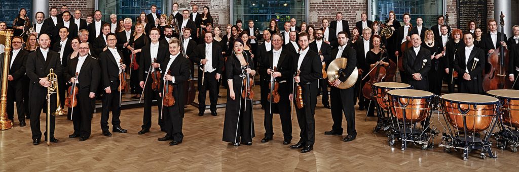 Members of the London Symphony Orchestra dressed in black tie apparel stand with their instruments.