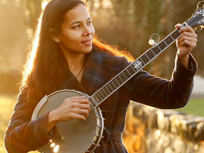 A woman tunes a banjo in a sunlit field.