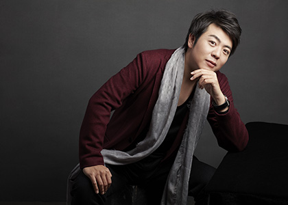 Pianist Lang Lang sits at a piano bench wearing a red blazer and grey scarf.