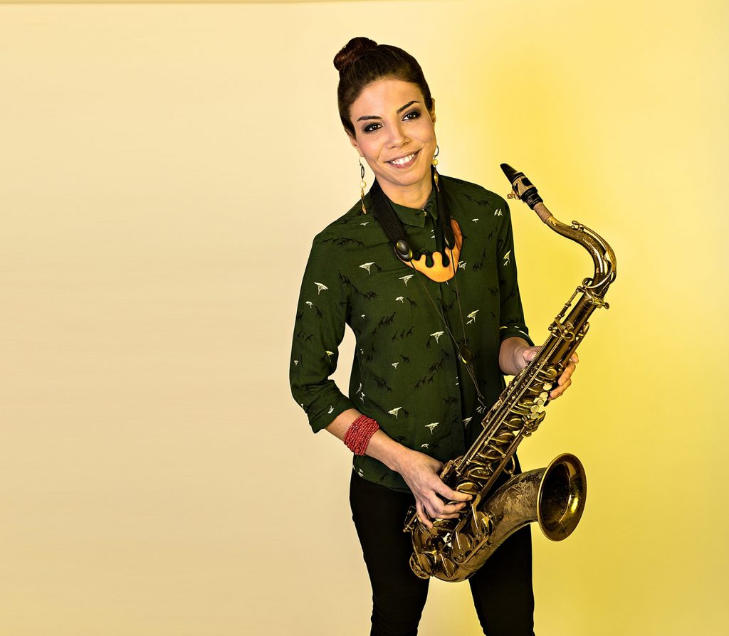 Saxophonist Melissa Aldana stands in front of a yellow background with her saxophone in her hands.