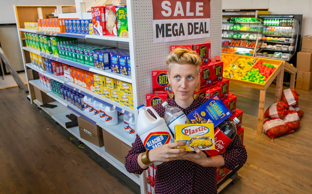 Woman with blonde hair stands with arms full of grocery items in what looks like a convenience store.