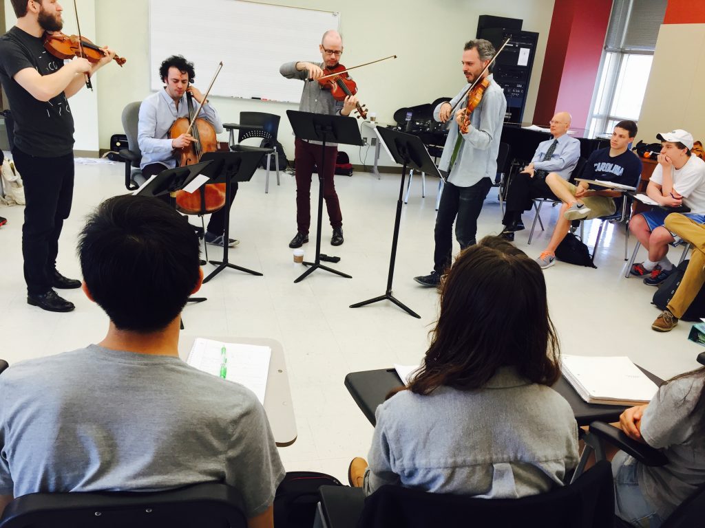 String quartet standing at music stands in a classroom, with seated students watching them play
