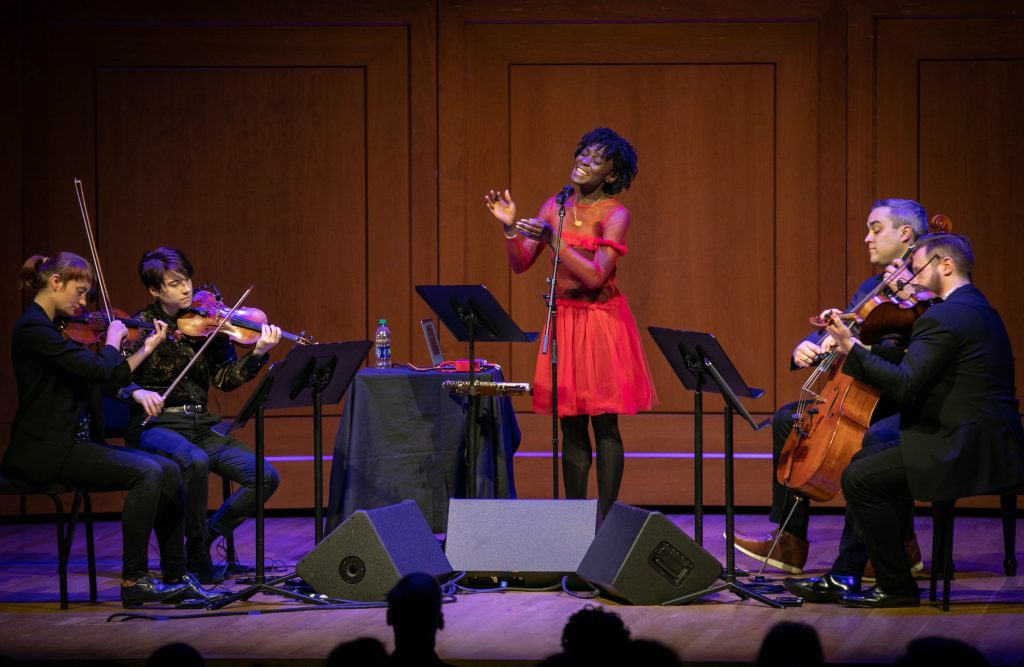 A Black woman in a red dress sings on a stage with a string quartet