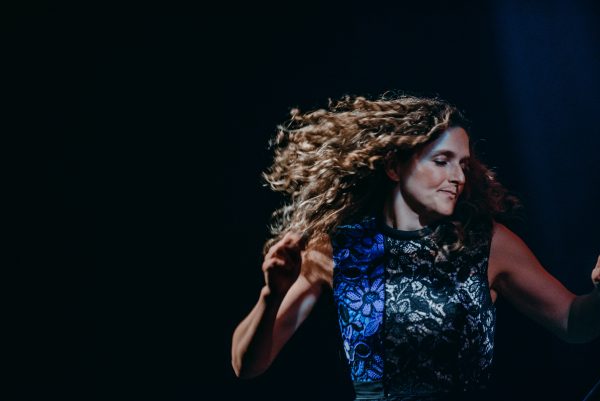 A white woman looking to the side, mid-twirl, with curly light brown hair and a lace dress against a dark background.