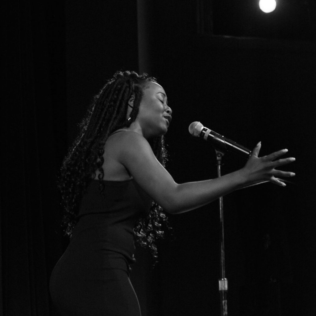 black and white image of a young Black woman with braids and a dark dress singing into a microphone