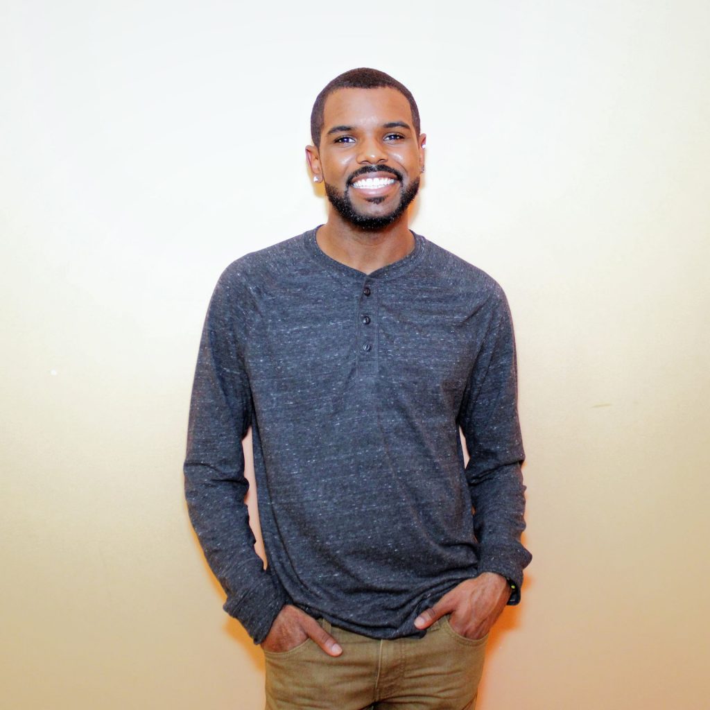 Young Black man wearing a dark blue henley long-sleeved shirt and beige pants smiling into camera
