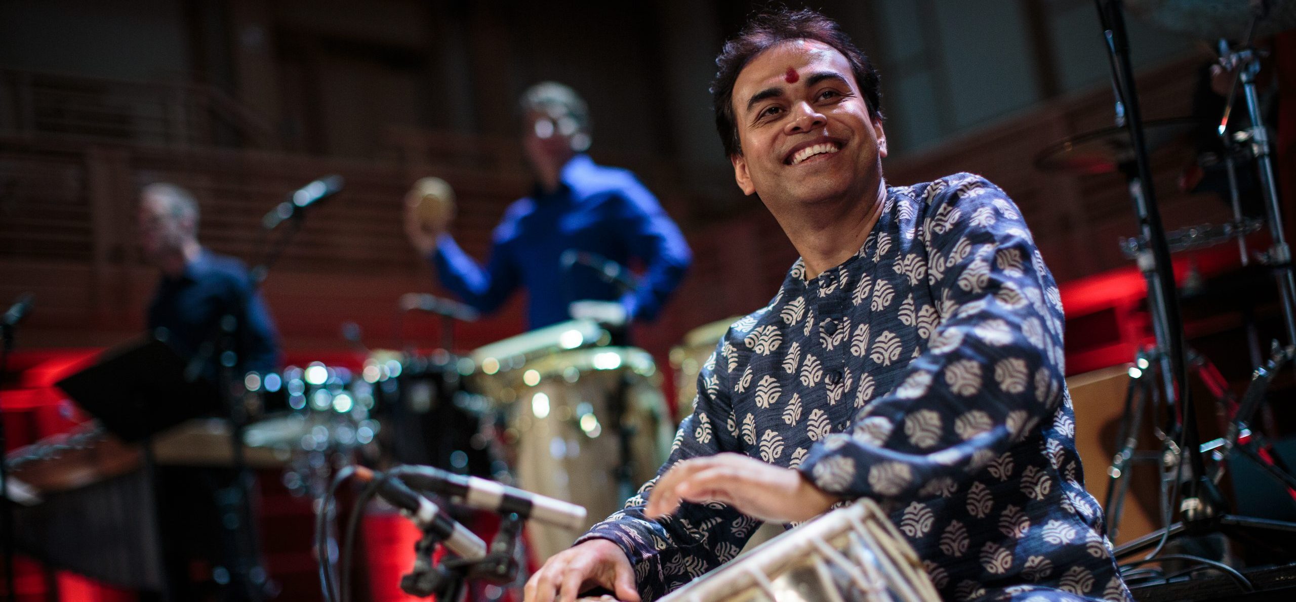 Sandeep Das, an Indian man wearing a blue and white patterned shirt, sits while playing the table, a drum