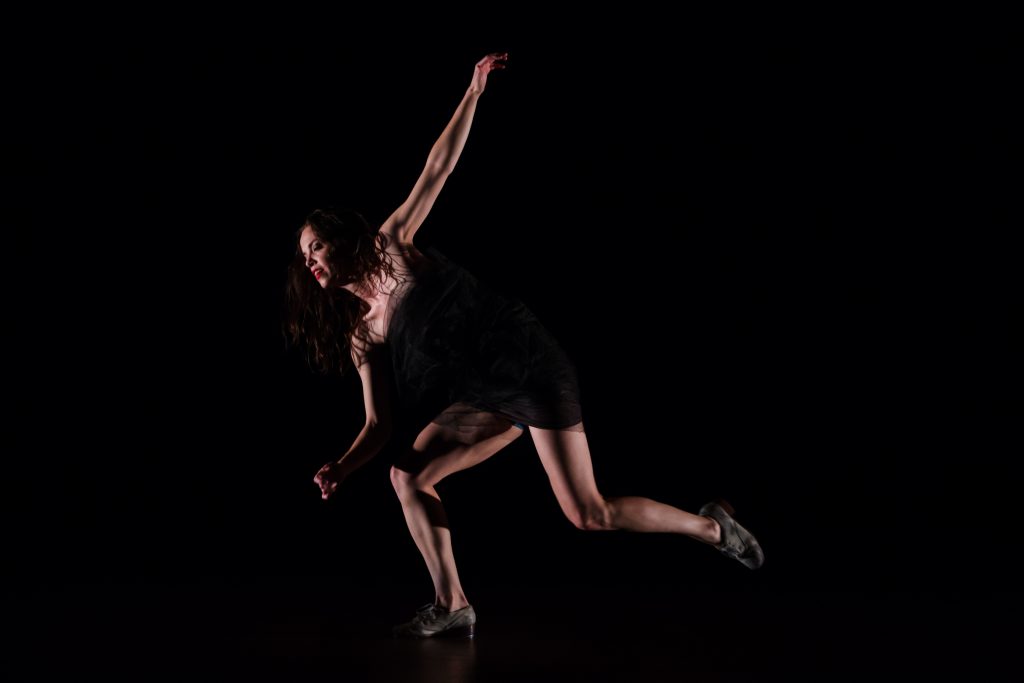 Michelle Dorrance, a white woman wearing a black leotard, tap dances on a darkly lit stage.