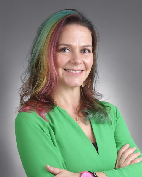 White woman with rainbow stripes in light brown hair wearing green v-neck top.