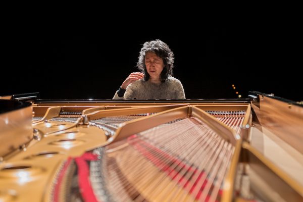Pianist Mitsuko Uchida, a Japanese woman, sits at a piano. The piano's lid is open, revealing the strings connected to the keys.