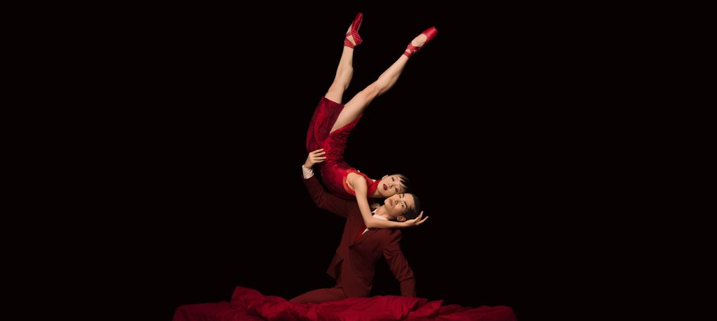 A male dancer, sitting on a red, velvet surface, holds a female dancer in the air. Both dancers are wearing red.
