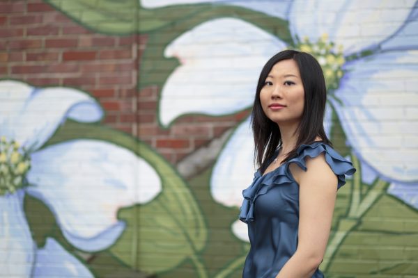 Clara Yang, an East Asian woman with long dark hair, wearing a sleeveless cornflower blue dress with ruffles and a necklace. She carries a neutral facial expression and is looking at the camera. She is standing in front of a mural depicting white flowers painted on a brick wall. The scene has bright and even lighting.