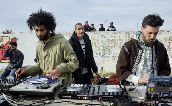 DJs stands behind turntables. They're outside. In the background, a man walks behind them. Graffiti can be seen behind them as well.
