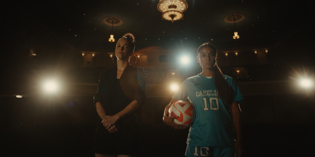 Two women stand side-by-side in an auditorium. One is dressed in dance gear. One is wearing a soccer uniform and holding a soccer ball.